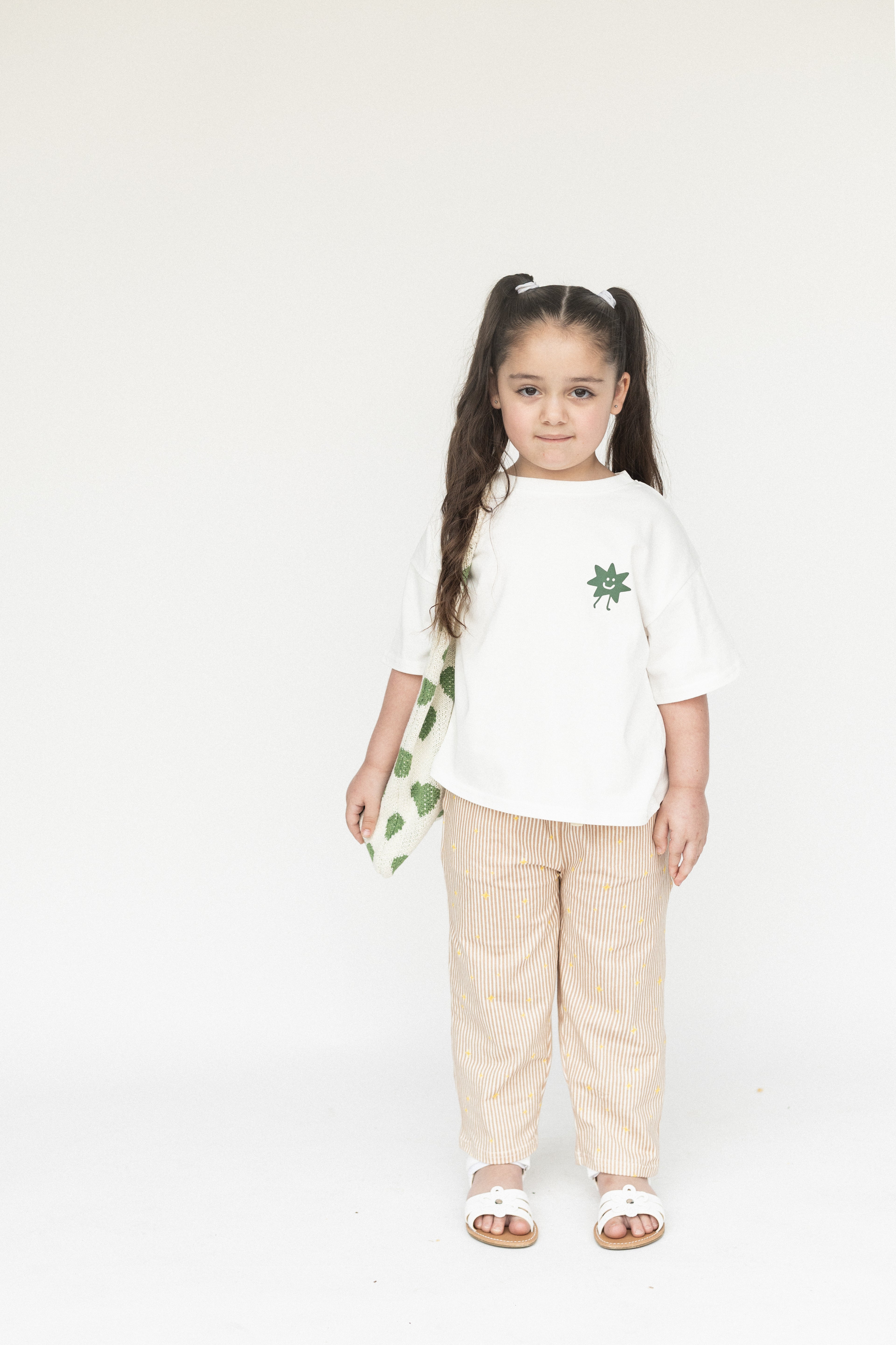 Young girl wearing a white t-shirt with green designs and beige pants on a white background