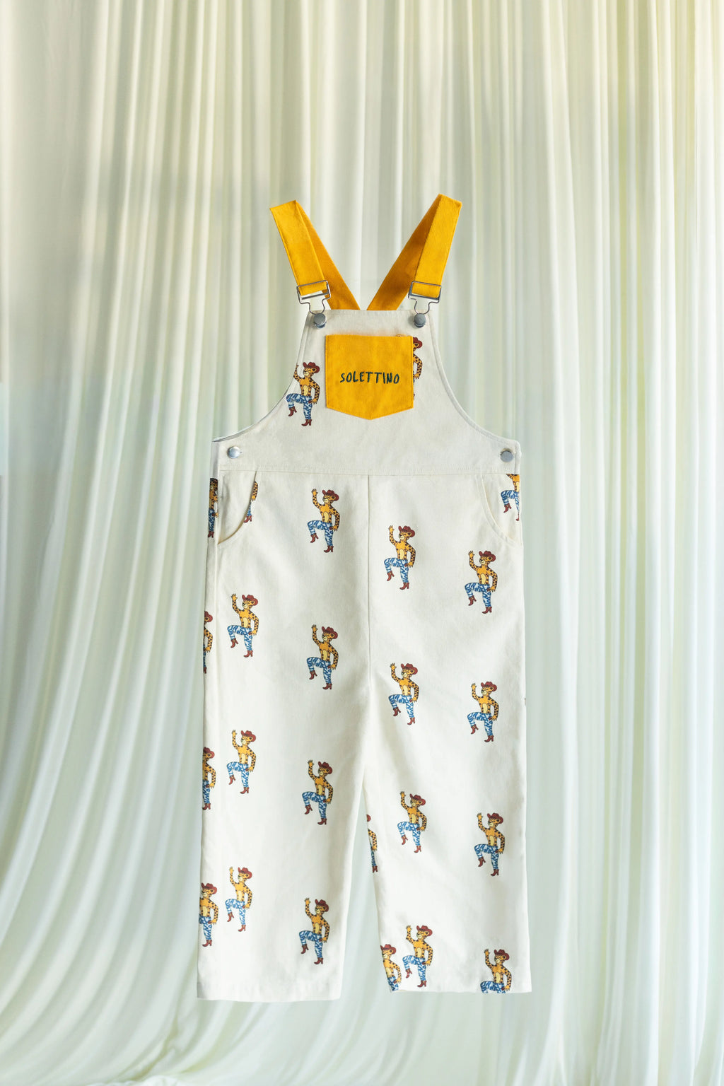 White overalls with cartoon character print and yellow straps on a light curtain background