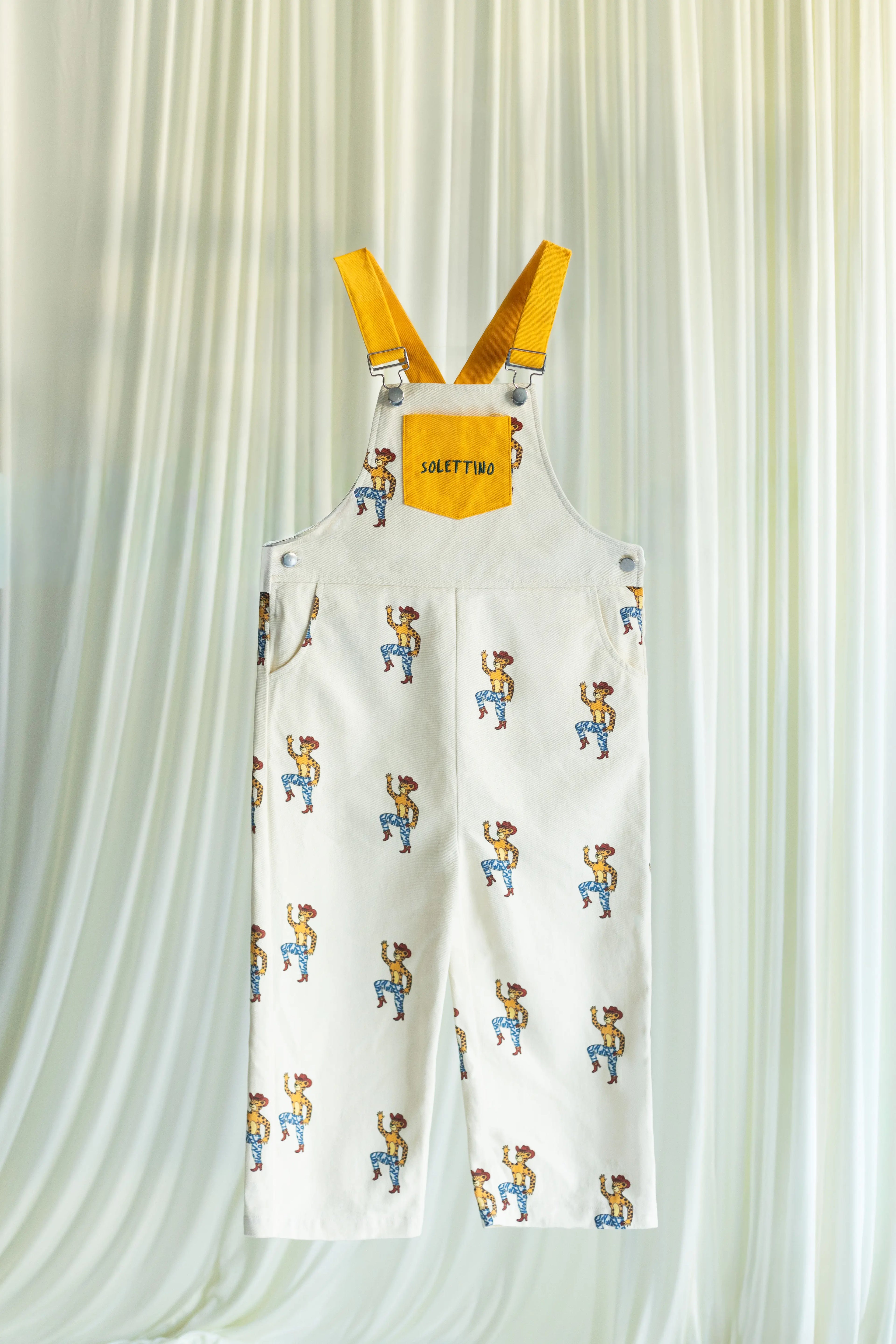 White overalls with cartoon character print and yellow straps on a light curtain background