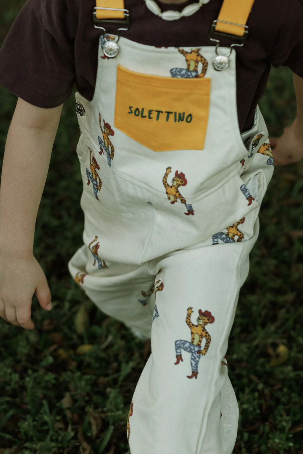 Child wearing white overalls with cartoon designs and a yellow pocket, standing on grass.