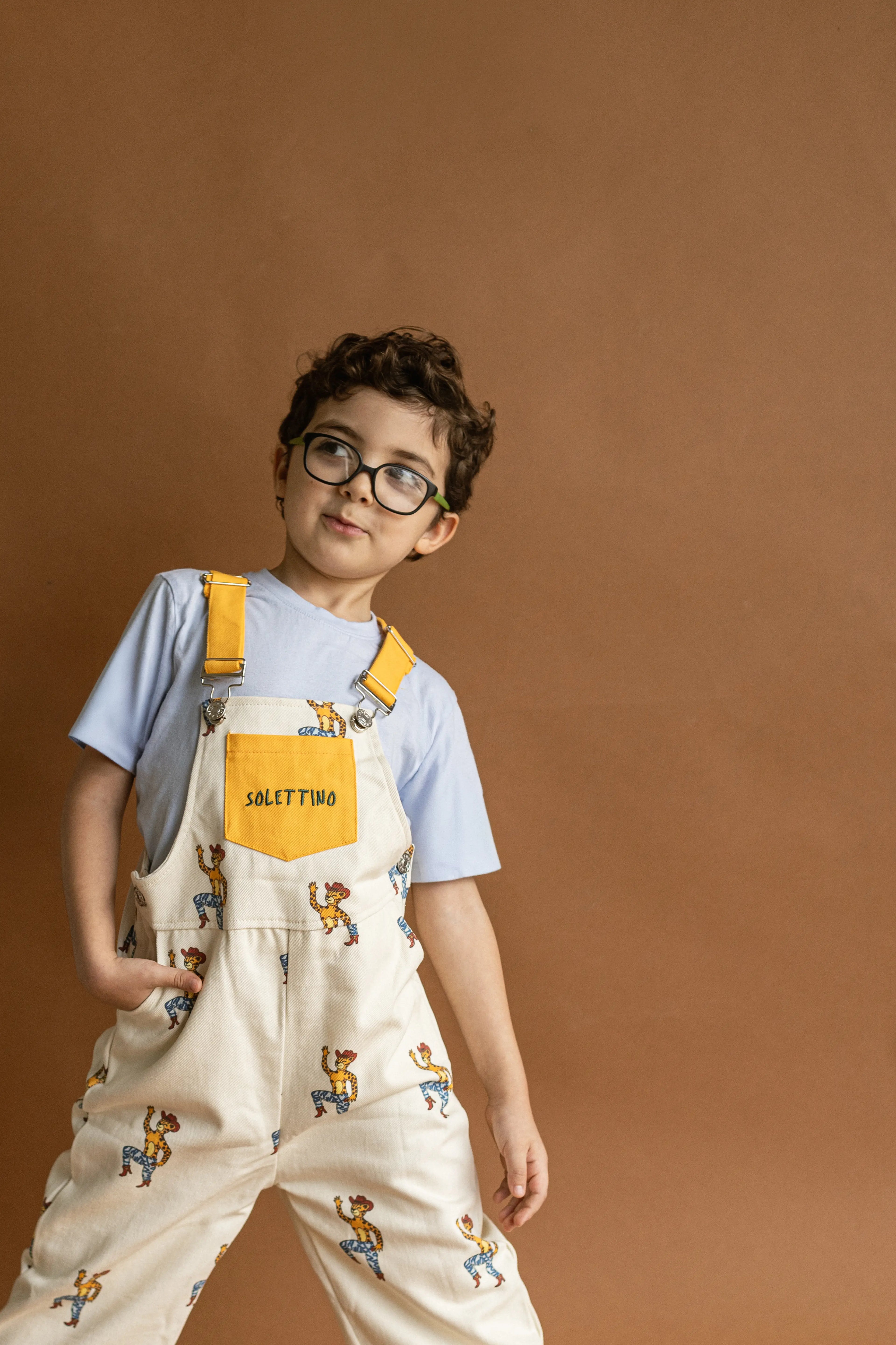Child wearing a patterned overalls and glasses against a brown background