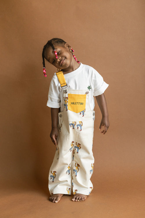 Child wearing a white t-shirt with colorful graphics and patterned overalls on a brown background