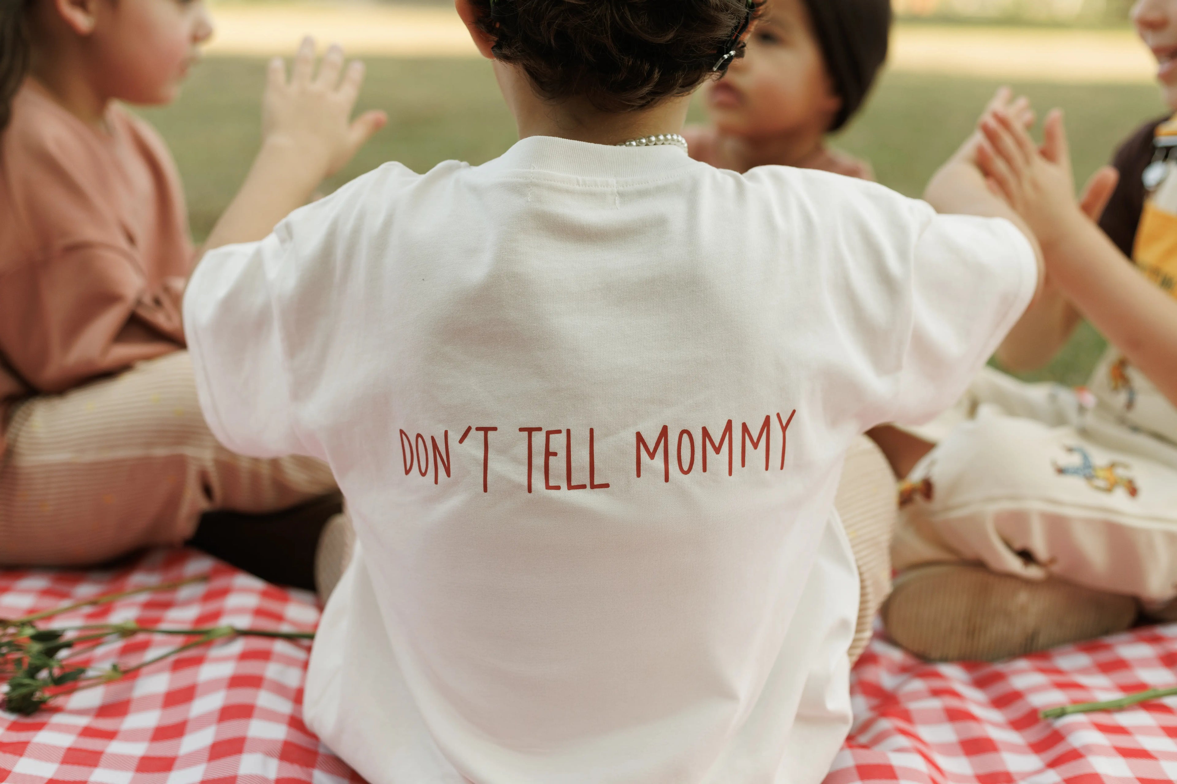 Child wearing a white t-shirt with 'DON'T TELL MOMMY' text at a picnic.
