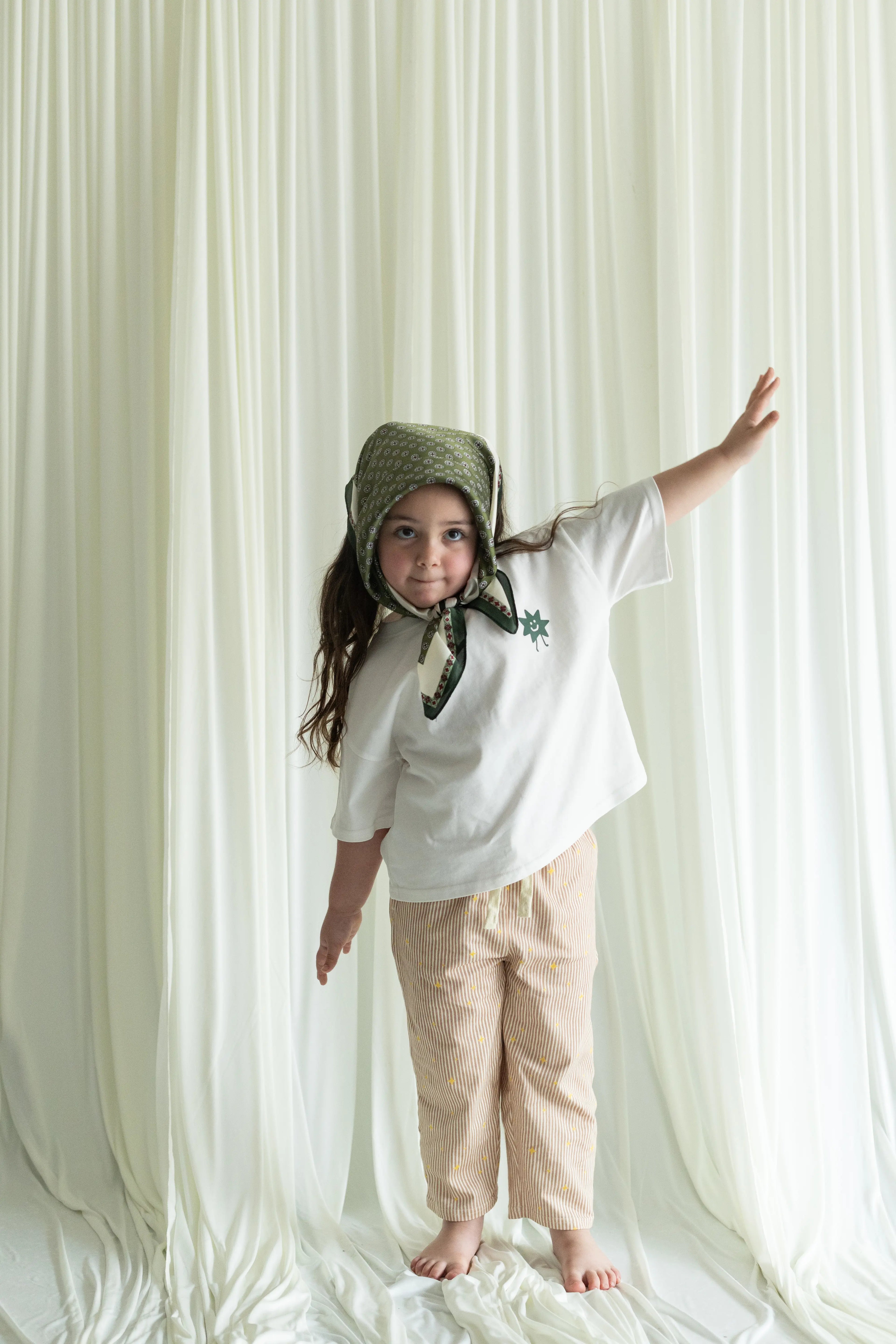 Child wearing a white shirt and beige pants with a green bonnet against a white curtain background
