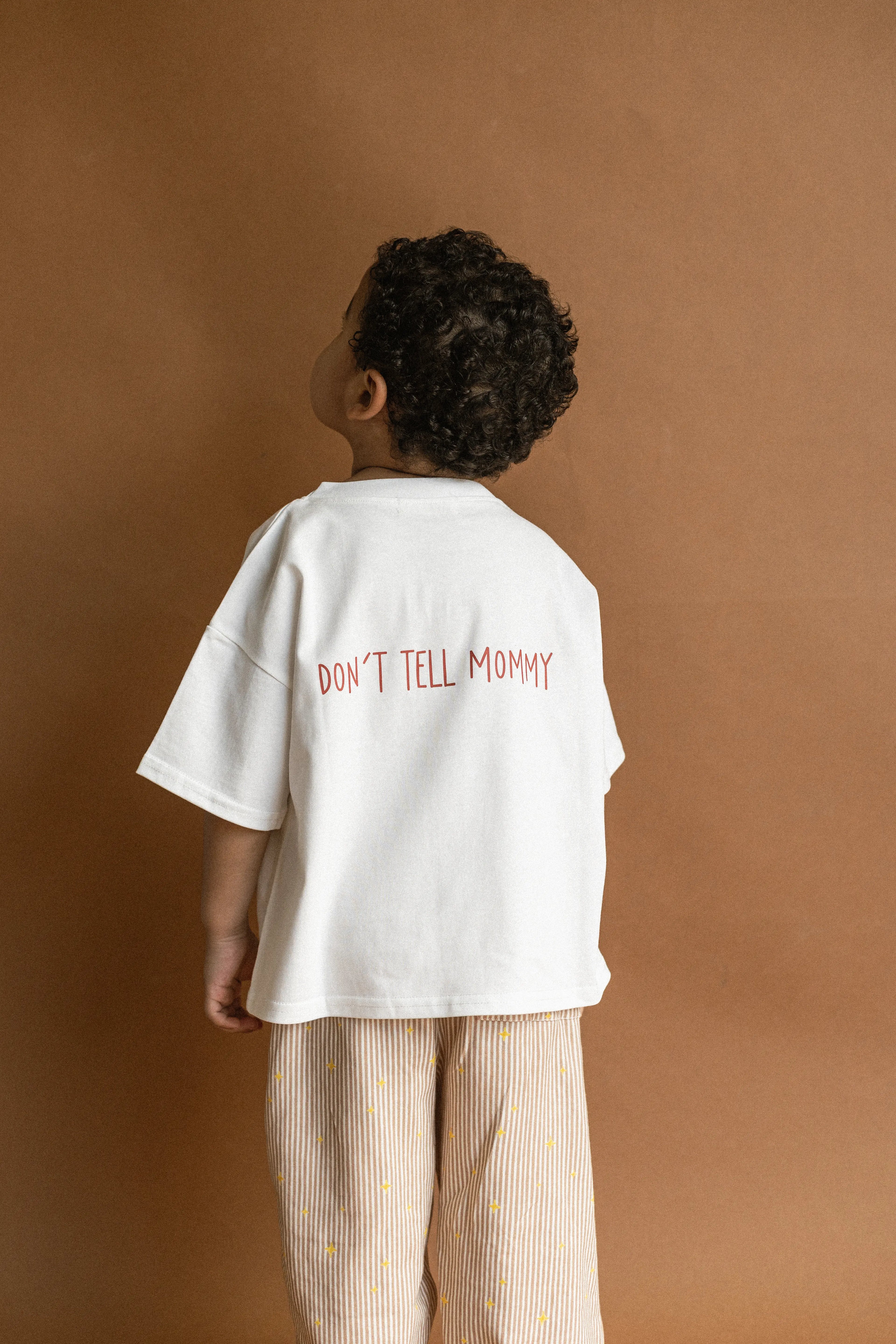Child wearing a white t-shirt with 'DON'T TELL MOMMY' text on a brown background