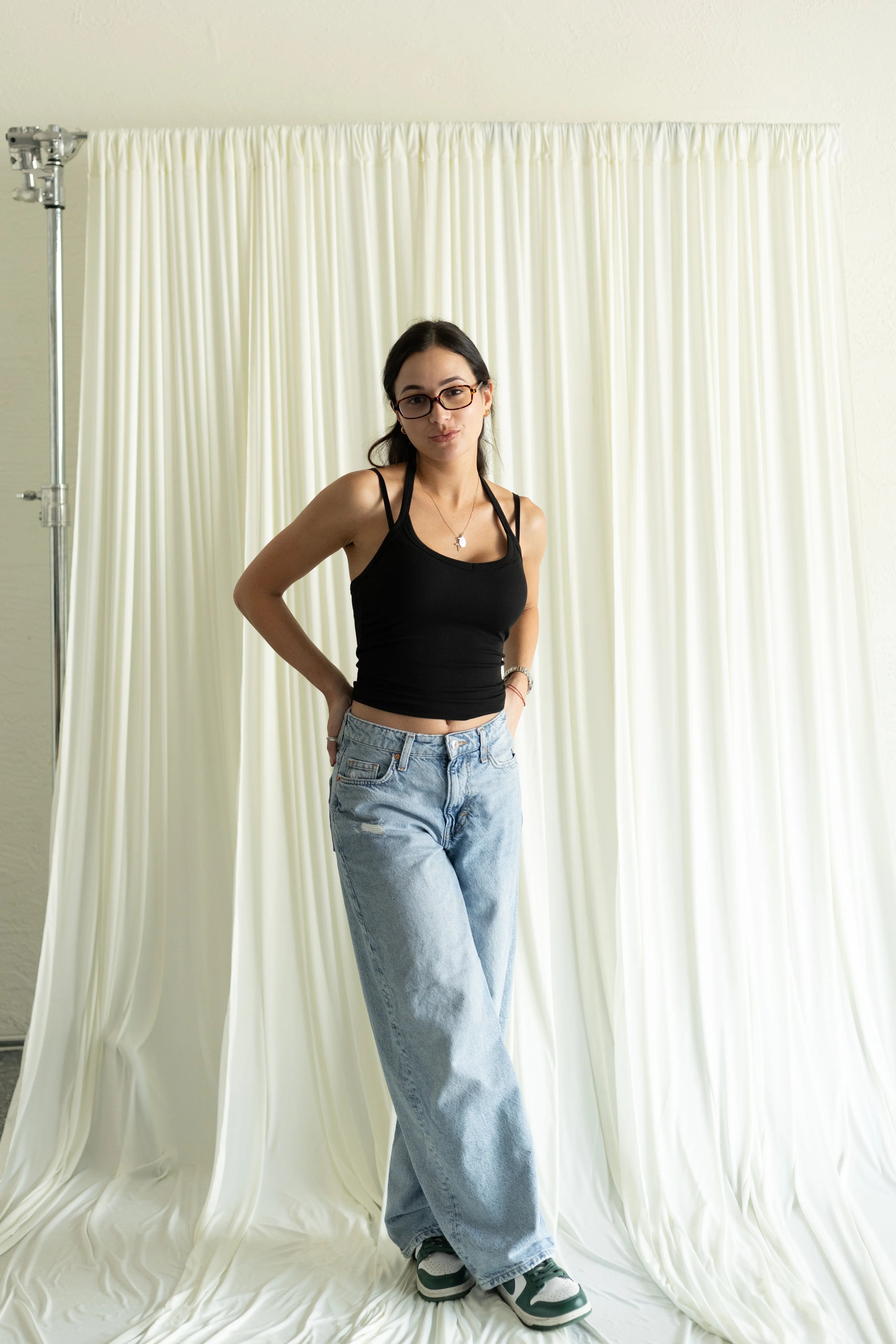 Woman wearing a black tank top and light blue jeans standing in front of white curtains.
